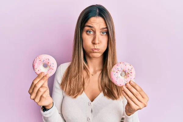 Brunette young woman holding tasty pink doughnuts puffing cheeks with funny face. mouth inflated with air, catching air.