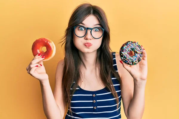 Young beautiful caucasian girl holding tasty colorful doughnuts puffing cheeks with funny face. mouth inflated with air, catching air.
