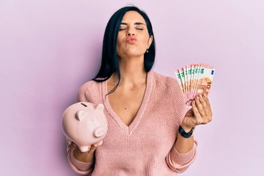 Avro banknotları tutan beyaz bir kadın ve kameraya bakan bir kumbara sevimli ve seksi bir şekilde öpüşüyor. aşk ifadesi. 