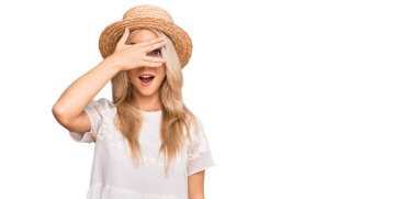 Yazlık şapkalı genç bir kız. Şoka girmiş, yüzünü ve gözlerini kapamış, utangaç ifadesiyle parmaklarının arasından bakıyor.. 