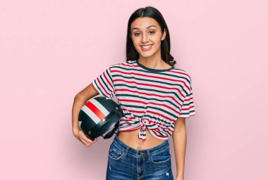 Motosiklet kaskı tutan genç İspanyol kız pozitif ve mutlu görünüyor. Kendine güvenen bir gülümsemeyle ayakta duruyor ve dişlerini gösteriyor. 