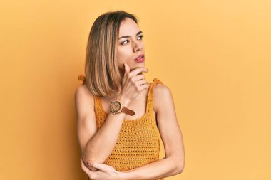 Kafkasyalı sarışın kadın, sıradan sarı tişörtler giyiyor, elleri çenesinde soru sormayı düşünüyor, düşünceli bir ifade. Düşünceli bir yüzle gülümsüyor. şüphe kavramı. 