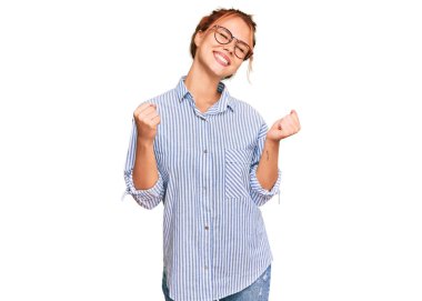 Genç kızıl saçlı, günlük kıyafetler ve gözlük takan mutlu ve heyecanlı bir şekilde kolları havada, gülümseyen ve başarı için bağıran bir jest yapıyor. kutlama konsepti. 