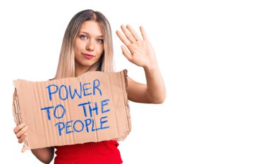 Genç, güzel, sarışın bir kadın. İnsanların afişini tutuyor. Eli açık, dur işareti yapıyor. 