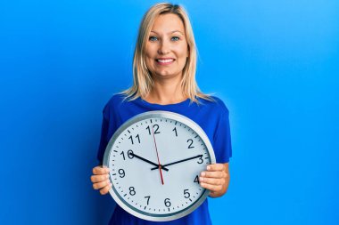 Beautiful middle age blonde woman holding big clock smiling with a happy and cool smile on face. showing teeth. 