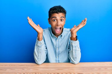 Genç ve yakışıklı Afro-Amerikan adam, gündelik giysiler içinde masaya oturmuş çılgın ve hayrete düşmüş bir şekilde başarıyı kollarını kaldırmış ve gözleri açık bir şekilde heyecanla haykırıyor. Kazanan konsept 