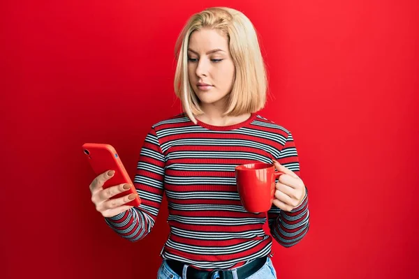Młoda Blondynka Używająca Smartfona Pijąca Filiżankę Kawy Zrelaksowana Poważnym Wyrazem — Zdjęcie stockowe