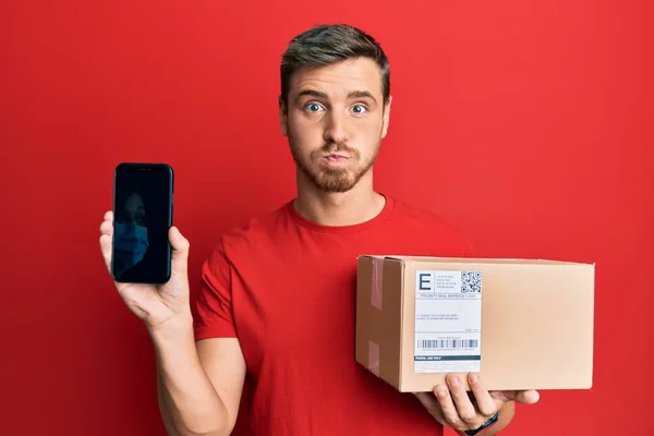 Handsome Caucasian Man Holding Delivery Package Showing Smartphone Screen Puffing — Stock Photo, Image