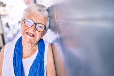 Yaşlı, yaşlı, kır saçlı, gülümseyen, açık havada duvara yaslanan yaşlı bir kadın.