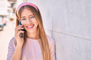 Kafkasyalı genç kız gülümsüyor. Duvara yaslanmış akıllı telefondan konuşuyor..