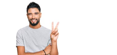 Gri bir tişört giyen sakallı genç adam kameraya bakıp gülümsüyor ve zafer işareti yapıyor. İki numara.. 