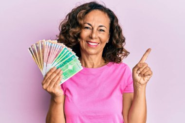 Orta yaşlı İspanyol kadın elinde Norveç kronu banknotları tutarken mutlu bir şekilde elini ve parmağını kenarı işaret ediyor. 