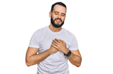 Sakallı, sıradan beyaz tişörtlü, elleri göğsünde, gözleri kapalı ve yüzünde minnet ifadesi olan genç bir adam. sağlık kavramı. 