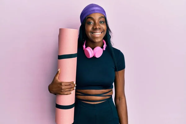 穿着运动服 手持瑜伽垫的年轻的非洲裔美国女人 站在那里看起来积极而快乐 脸上带着自信的笑容 露出牙齿 — 图库照片