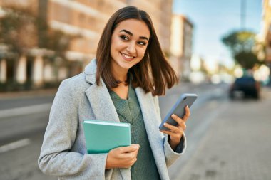 Genç İspanyol kız mutlu bir şekilde gülümsüyor. Akıllı telefon kullanıyor ve şehirde kitap tutuyor..