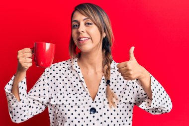 Genç ve güzel bir kadın bir kupa kahve içiyor mutlu ve pozitif gülümsüyor, başparmağıyla mükemmel ve onay işareti yapıyor. 