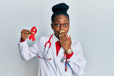 Genç Afro-Amerikan kadın doktor üniforması giyiyor. Elinde kırmızı kurdele var. Ağzı elle kapatıyor. şaşırmış ifade 