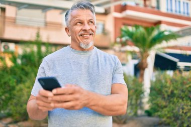 Orta yaşlı, ispanyol, kır saçlı, mutlu gülümseyen bir adam parkta akıllı telefon kullanıyor..