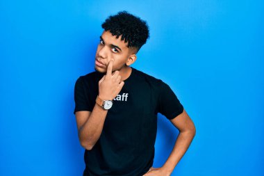 Genç, Afro-Amerikalı, personel tişörtü giymiş. Gözünü dikip jestini izliyor. Şüpheli bir ifade. 
