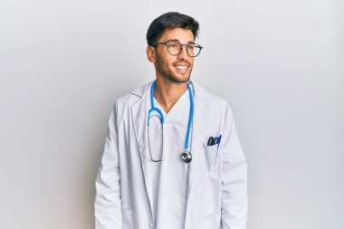 Doktor üniforması ve steteskop giymiş genç yakışıklı bir adam yüzünde gülümseme, doğal bir ifade ile yan yana bakıyor. Kendinden emin gülen. 