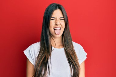 Genç esmer kadın kırmızı arka plan üzerine gündelik beyaz tişört giyip komik bir ifadeyle dilini dışarı çıkarıyor. duygu kavramı. 