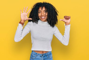 Genç Afro-Amerikan kızı gündelik giysiler giyip altı numaralı parmaklarıyla işaret ederken kendinden emin ve mutlu bir şekilde gülümsüyor.. 