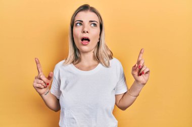 Gündelik beyaz tişört giyen güzel beyaz kadın şaşkın şaşkın yukarı bakıyor ve parmaklarıyla işaret ediyor ve kollarını kaldırıyor.. 
