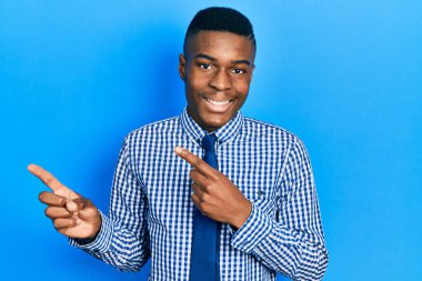 Genç Afro-Amerikan adam iş gömleği ve kravat takıyor gülümsüyor ve kameraya bakıyor iki eliyle ve parmaklarıyla kenarı işaret ediyor.. 