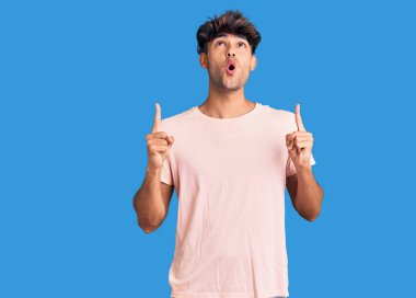 Gündelik giysiler giyen genç İspanyol adam şaşırmış ve şaşırmış bir şekilde yukarı bakıyor ve parmaklarıyla işaret ediyor ve kollarını kaldırıyor.. 