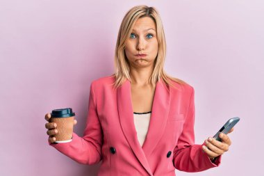 Güzel orta yaşlı sarışın kadın akıllı telefon kullanıyor ve komik suratlı kahve şişiren yanaklar içiyor. Ağzı havayla şişer, havayı yakalar.. 