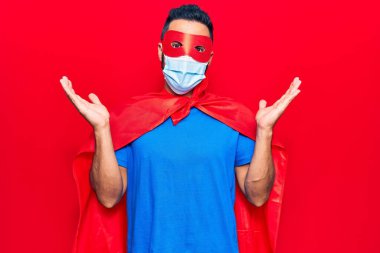 Young hispanic man wearing super hero costume and medical mask celebrating achievement with happy smile and winner expression with raised hand 