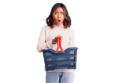 Genç, güzel, melez bir kadın elinde alışveriş sepeti tutarken ağzı açık kalmış, korkmuş ve şaşkın bir şekilde şaşırmış. 