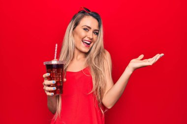 Güzel sarışın kadın kırmızı arka planda saman kullanarak kola içiyor. mutlu bir gülümsemeyle başarıyı kutluyor. ve kazanan el kaldırarak ifade ediyor.