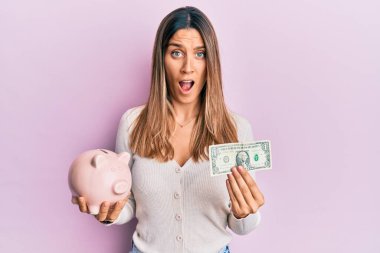 Elinde bir dolarlık banknot ve kumbara tutan esmer genç bir kadın şaşkınlık ve şaşkınlık dolu bir ifade, korku ve heyecanlı bir yüz ifadesi ile şok olmuştu.. 