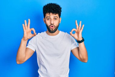 Sakallı, sıradan beyaz tişörtlü genç bir Arap. Şaşırmış ve şaşırmış görünüyor. Onaylama sembolünü parmaklarıyla yapıyor. çılgın ifade 