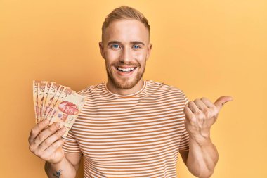 Meksika Pezosu 'nu elinde tutan genç, beyaz bir adam başparmağını yana kaldırmış, ağzı açık gülümsüyor. 