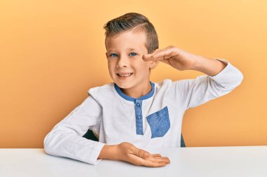 Sevimli beyaz çocuk, sıradan kıyafetler giyip masaya oturmuş büyük ve büyük boyut işareti, ölçü sembolü göstererek el kol hareketi yapıyor. Kameraya bakıp gülümsüyor. ölçüm kavramı. 