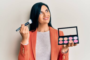 Makyaj fırçası ve allık paleti tutan genç beyaz kadın gülümsüyor ve başka tarafa bakıyor.. 