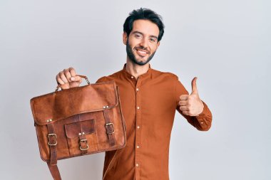 Deri çantalı genç adam mutlu ve pozitif gülümsüyor, başparmağıyla mükemmel ve onay işareti yapıyor. 