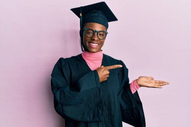 Young african american girl wearing graduation cap and ceremony robe amazed and smiling to the camera while presenting with hand and pointing with finger. 