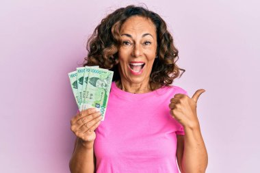 Orta yaşlı İspanyol kadın elinde 10000 Güney Koreli para kazandı. Parmağını yan tarafa kaldırdı ve ağzı açık gülümsüyor. 