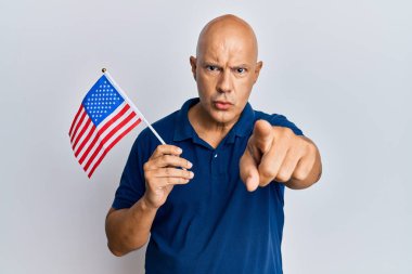 Orta yaşlı kel bir adam, elinde ABD bayraklarıyla kamerayı işaret ediyor. Kendine güvenen bir jest, ciddi görünüyor. 