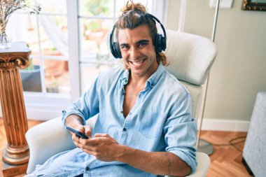 Genç İspanyol adam evde akıllı telefon ve kulaklık kullanarak mutlu bir şekilde gülüyor.