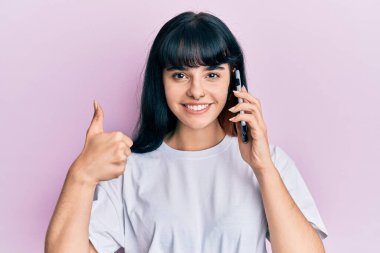 Genç İspanyol kız akıllı telefondan konuşuyor mutlu ve olumlu gülümsüyor, başparmağıyla mükemmel ve onay işareti yapıyor. 