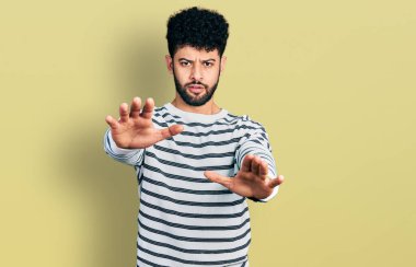 Sakallı, sıradan çizgili süveter giyen genç bir Arap. Avuç içi hareketlerini durduruyor. Öfkeli ve hayal kırıklığı dolu bir ifade takınıyor. 