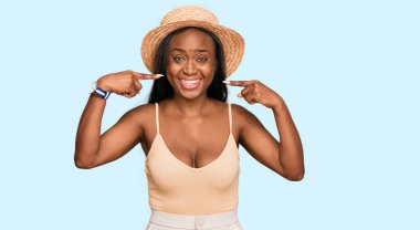 Yazlık şapkalı, neşeli gülümseyen, parmaklarıyla ve ağzıyla işaret eden genç siyahi bir kadın. diş sağlığı kavramı. 