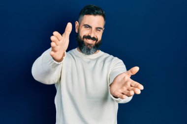 Gündelik giysiler giymiş genç İspanyol adam kameraya bakıp sarılmak için kollarını açmış gülümsüyor. Mutluluğu kucaklayan neşeli bir ifade.. 