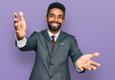 Genç Afro-Amerikalı iş kıyafetleri giymiş kameraya sarılıp gülümseyen genç bir adam. Mutluluğu kucaklayan neşeli bir ifade.. 