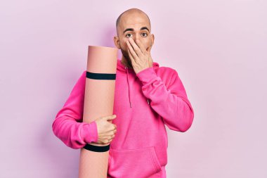 Elinde yoga minderi tutan genç kel bir adam ağzını eliyle kapatıyor, şok olmuş ve hata yapmaktan korkuyor. şaşırmış ifade 