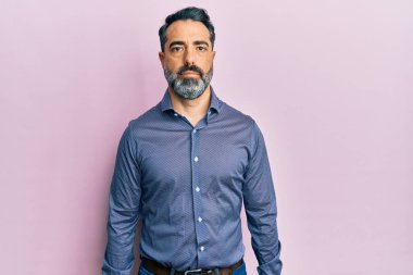 Sakallı, gri saçlı, iş kıyafetleri giyen orta yaşlı bir adam. Yüzünde ciddi bir ifadeyle rahatlamış. Basit ve doğal bir şekilde kameraya bakıyorsun.. 